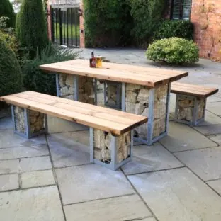 An outdoor rectangular dining table and bench seats made from wooden tops and rock filled metal gabion cage legs on a patio