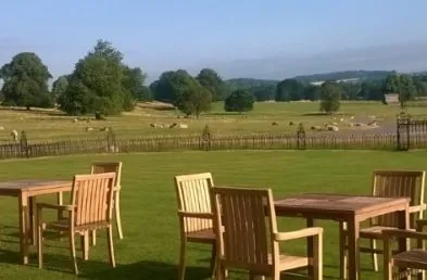 Three teak outdoor dining tables with four chairs around each one on a lawn overlooking a field of sheep