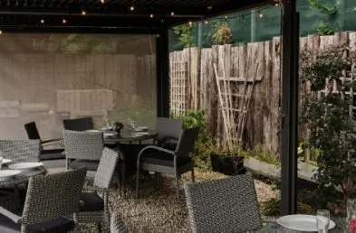 A close up of 3 round grey rattan tables and chairs sets underneath a grey metal gazebo with a flat roof on a gravel patio