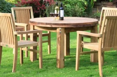 A round wooden table on a lawn with 4 teak stacking armchairs around it