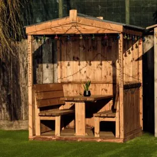 A wooden outdoor dining cabin with roof and back but with open half sides and an open frontage. Bench seats and table for 6 people are fixed inside