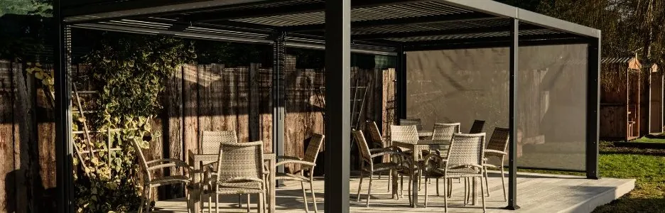 A grey framed rectangular metal gazebo with 3 sets of outdoor dining tables and chairs on a deck